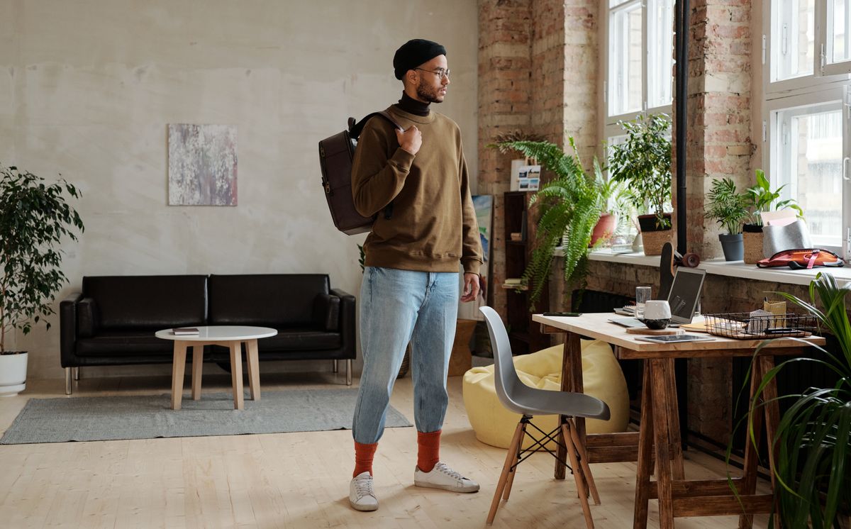 A man in knitwear and cuffed jeans carrying a backpack indoors.
